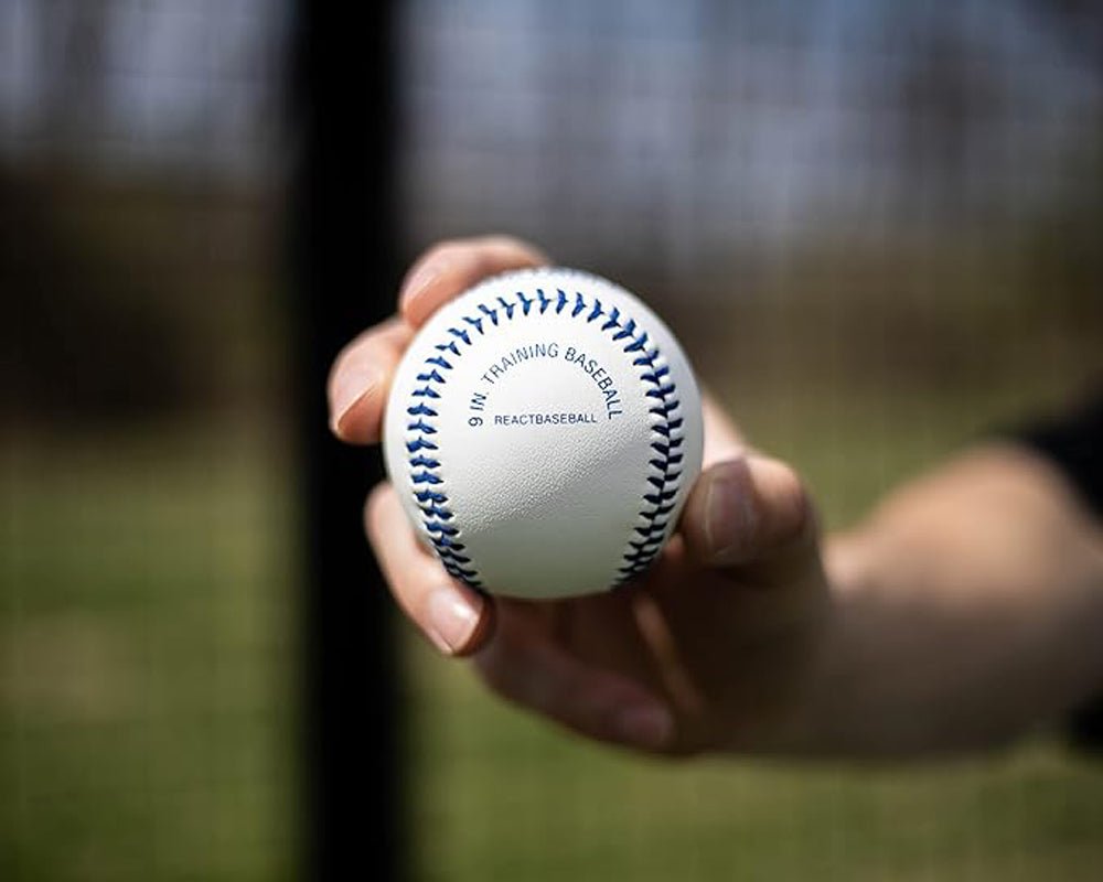 Hand holding a white training baseball with blue stitching against a blurred outdoor background