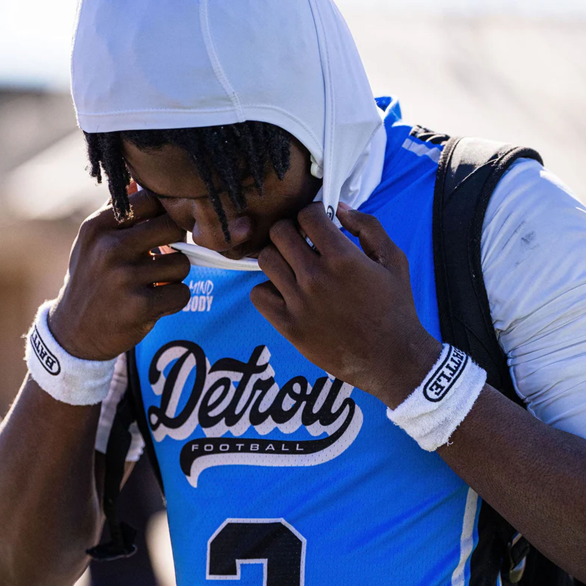 Person wearing a blue sports jersey and thick arm bands with 'Detroit Football' text, holding a white towel to their face.