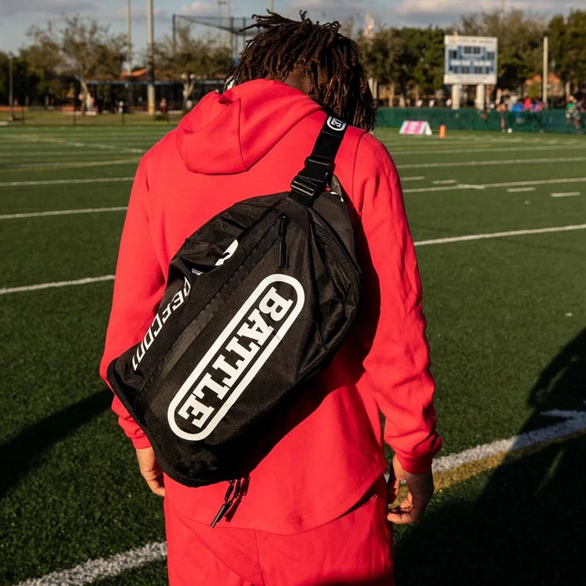 Person wearing a red jacket with a black sling bag 'BATTLE' backpack on a sports field.