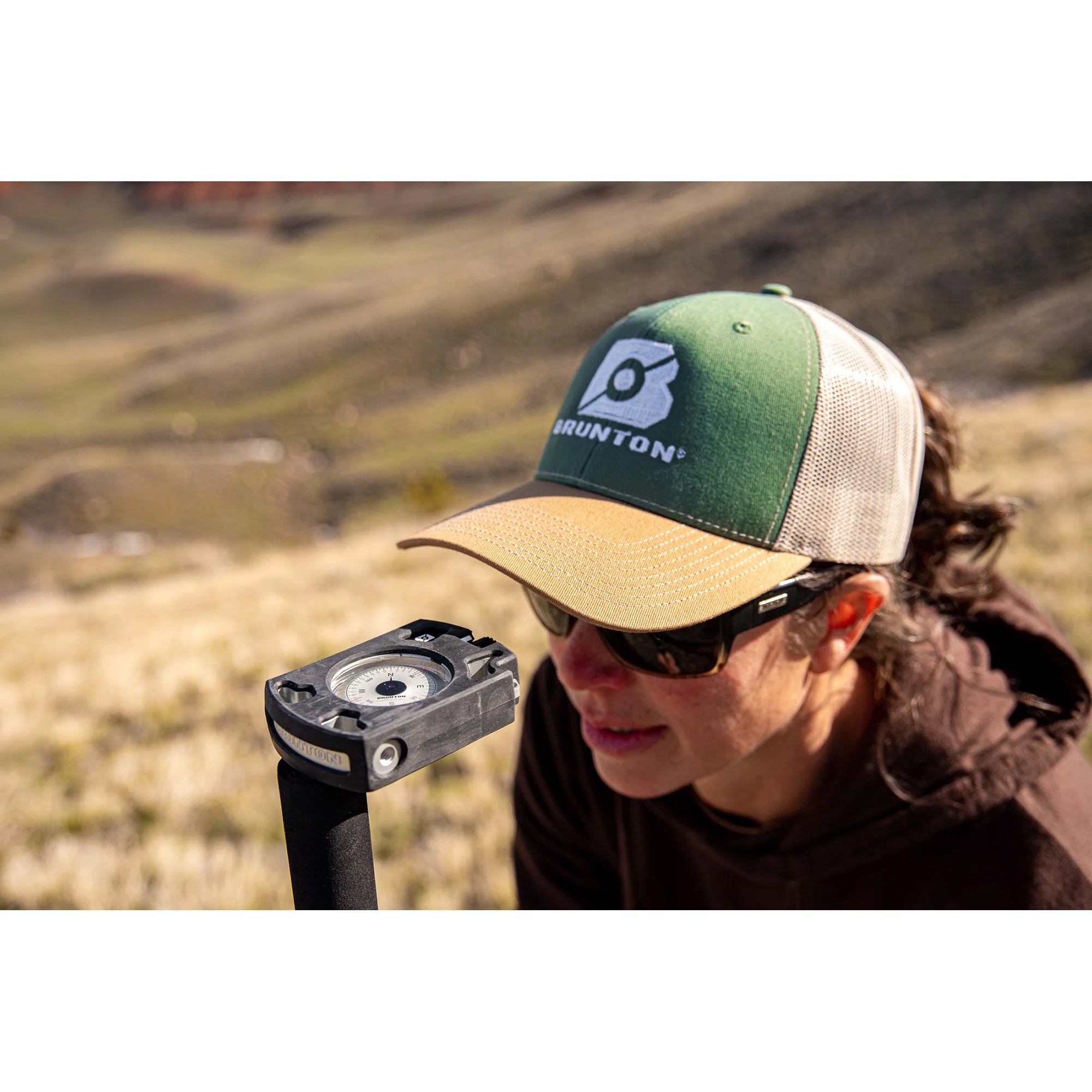Person wearing a Brunton cap with a Brunton tool in the foreground, set against a natural landscape.