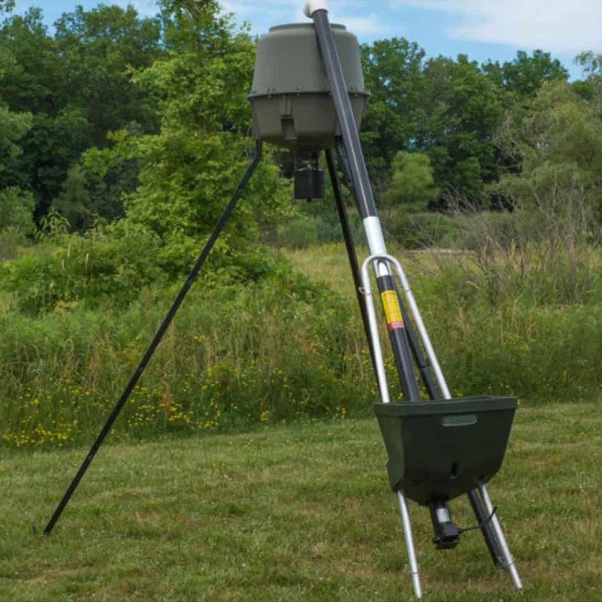 Hunting feeder filler on a tripod in a grassy field with trees in the background
