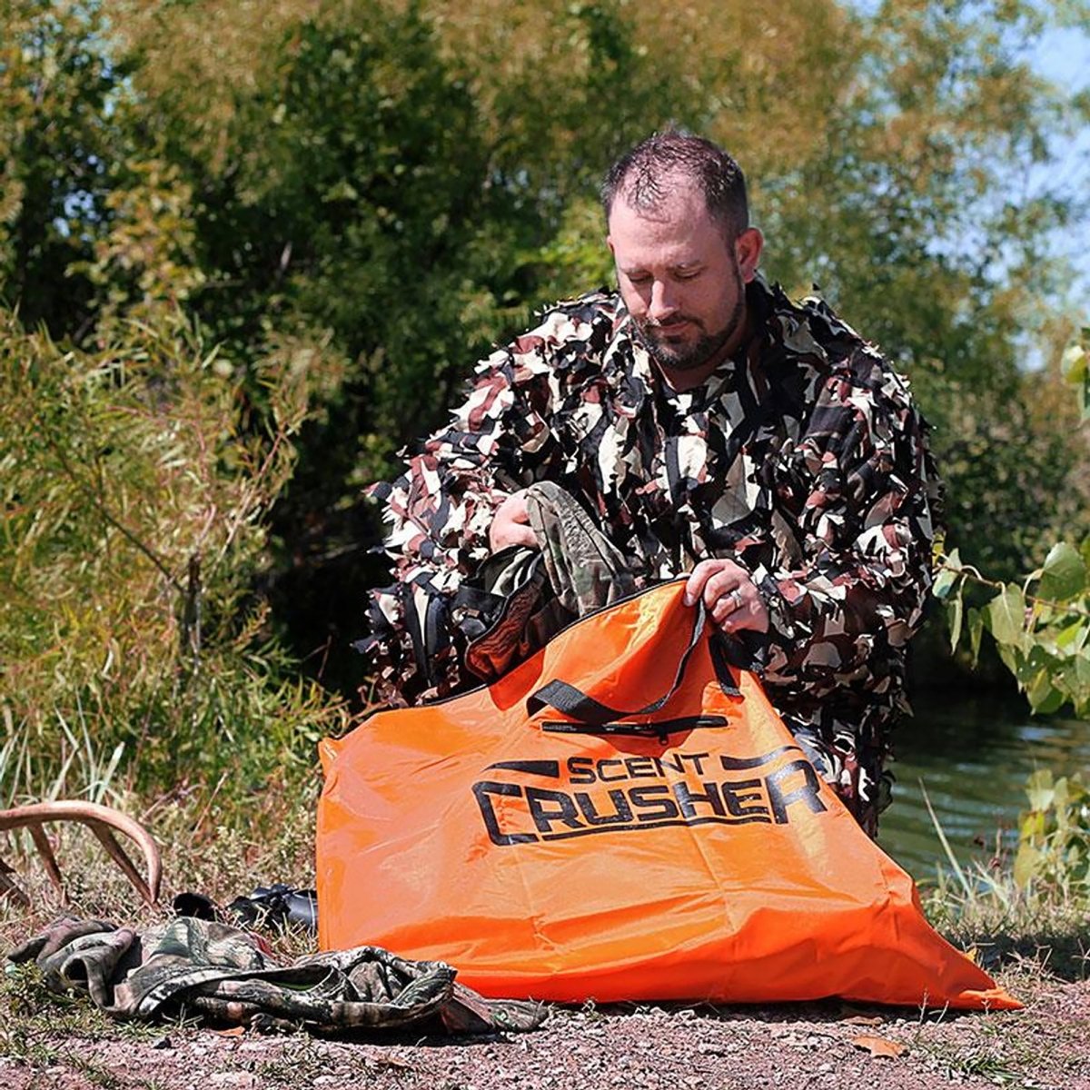 Man in camouflage clothing opening an orange 'Scent Crusher' bag outdoors.