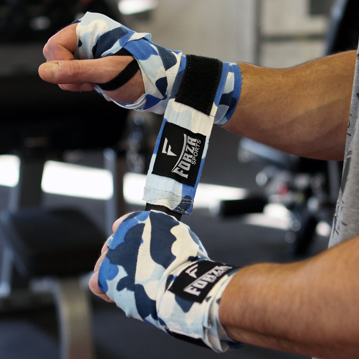 Hand wrapped in blue and white camouflage boxing tape with Forza brand logo, blurred gym background