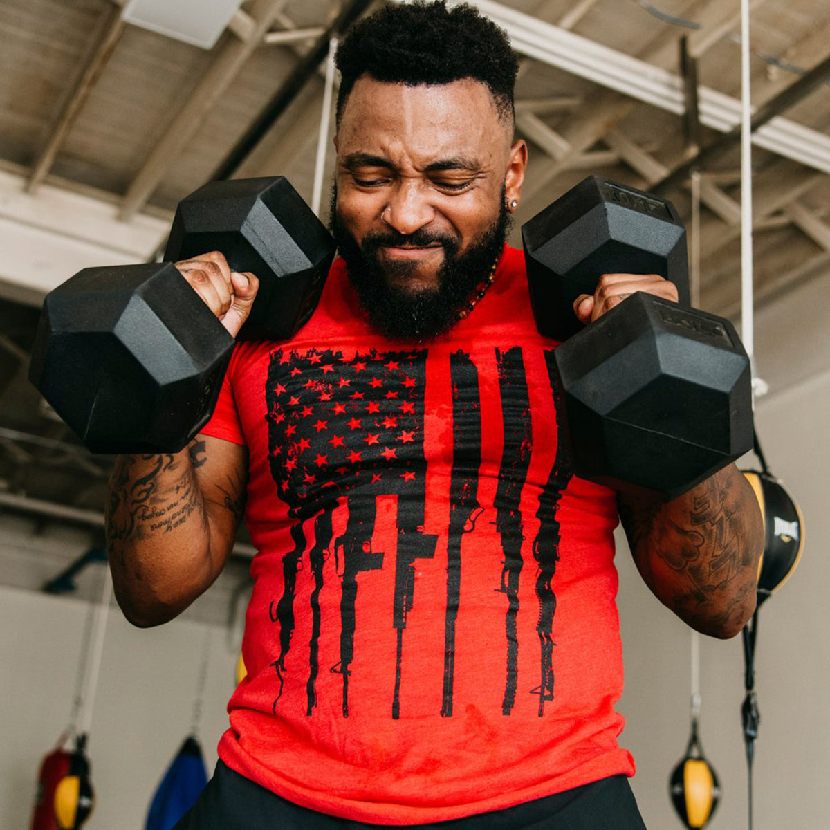 Man lifting black dumbbells wearing a red t-shirt with a black graphic design in a gym setting.