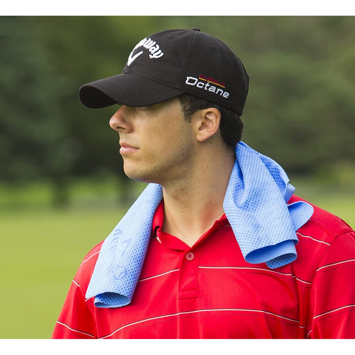 Man wearing a black cap with a logo and a red shirt, holding a blue golf cooling towel over his shoulder on a golf course.
