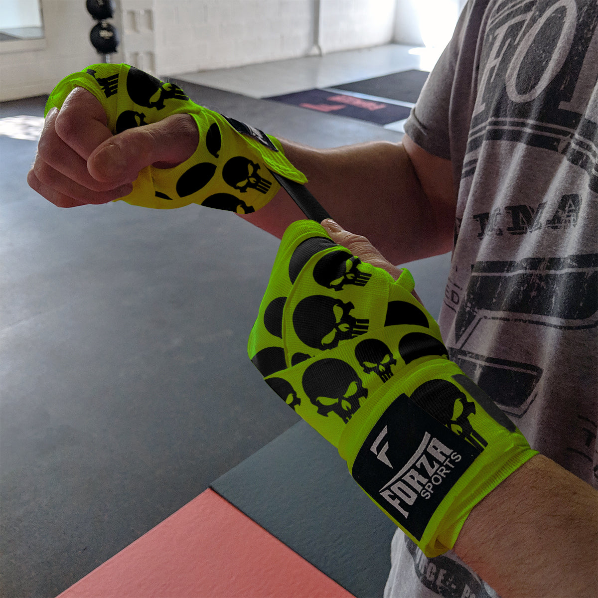 Person wearing bright green boxing wraps with black skull patterns, branded 'Forza Sports', in a gym setting.