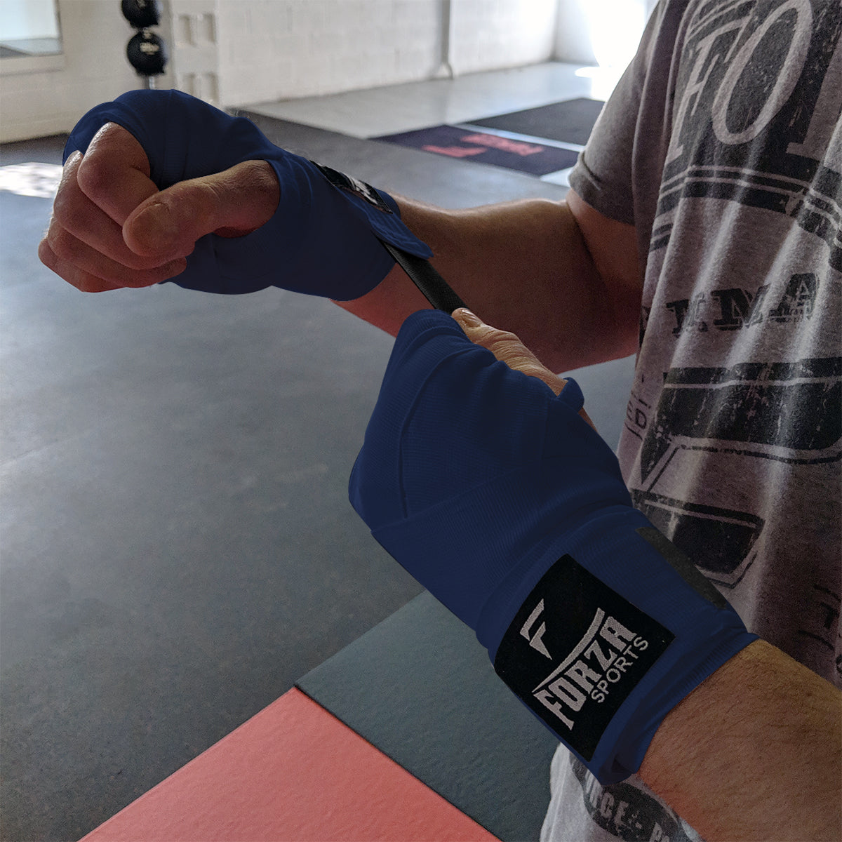 Person wrapping their hands with blue boxing hand wraps featuring a brand logo on a gym floor.