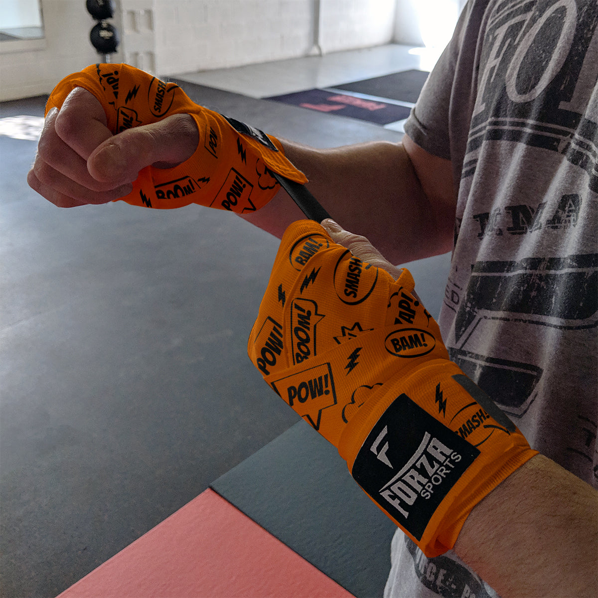 Person wearing orange boxing wraps with black text and logos on a gym floor.