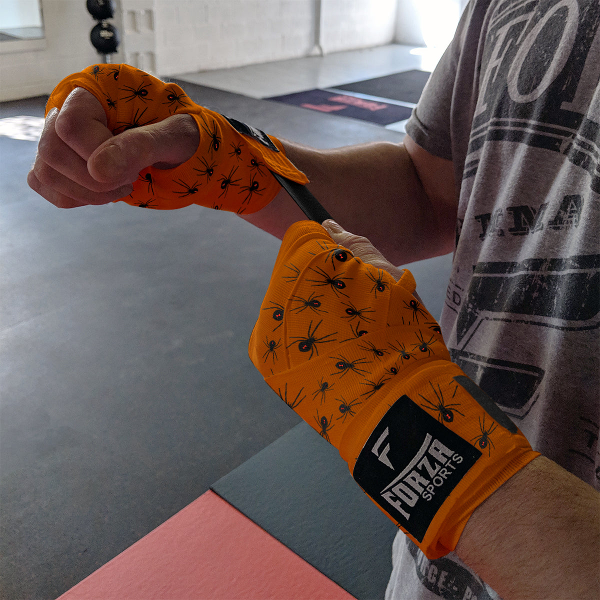 Person wearing orange boxing wraps with black spider design, featuring a visible brand logo, in an indoor setting.