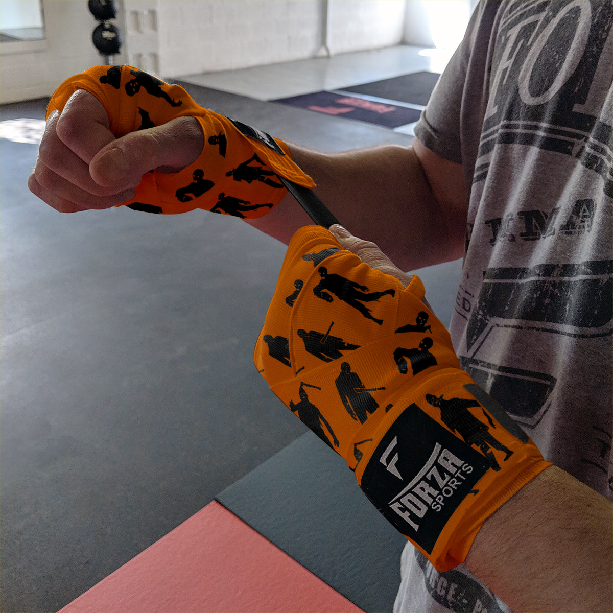 Person wrapping their wrist with orange boxing wraps featuring black silhouettes and 'Forza Sports' branding.