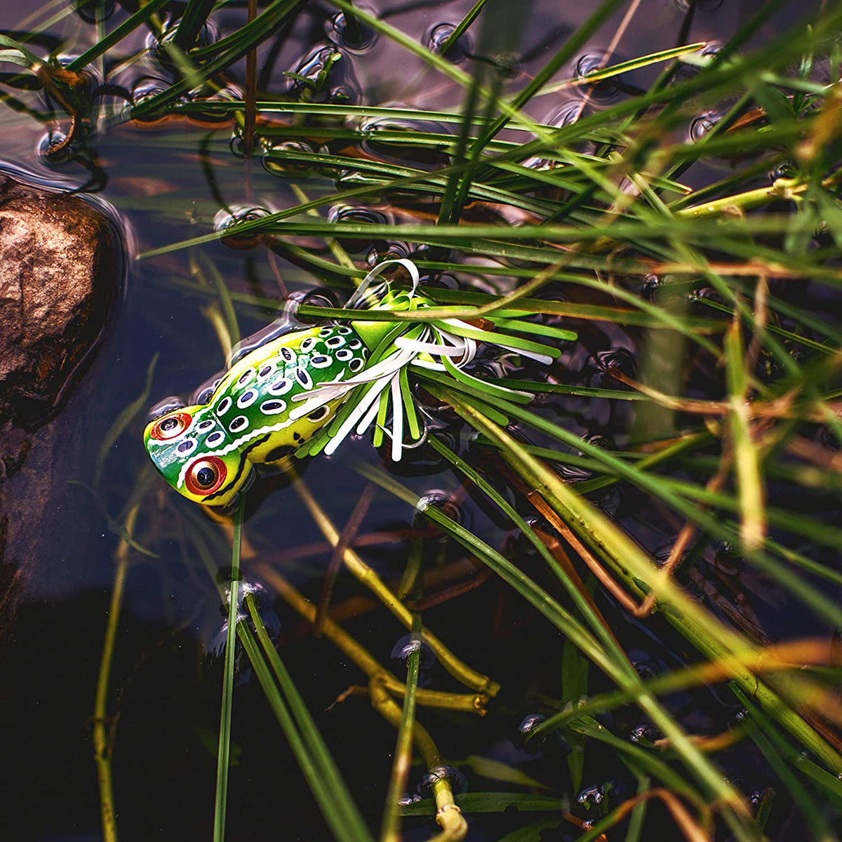 Fishing lure shaped like a frog floating on water with grasses