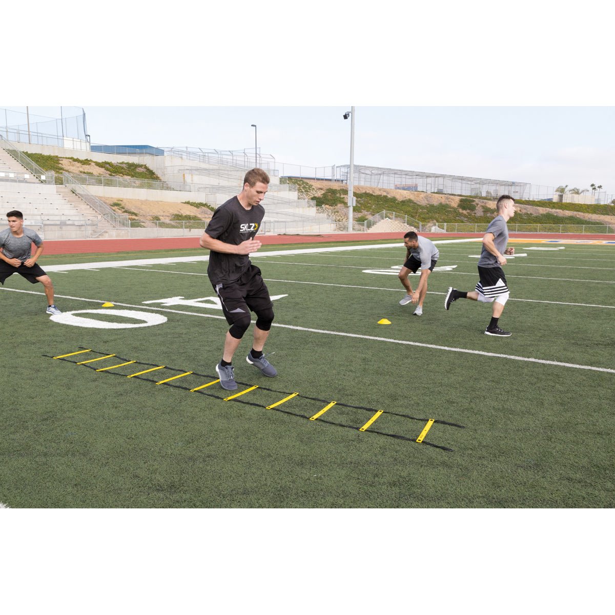 Person using a yellow agility quick ladder pro on a sports field with other people in the background.