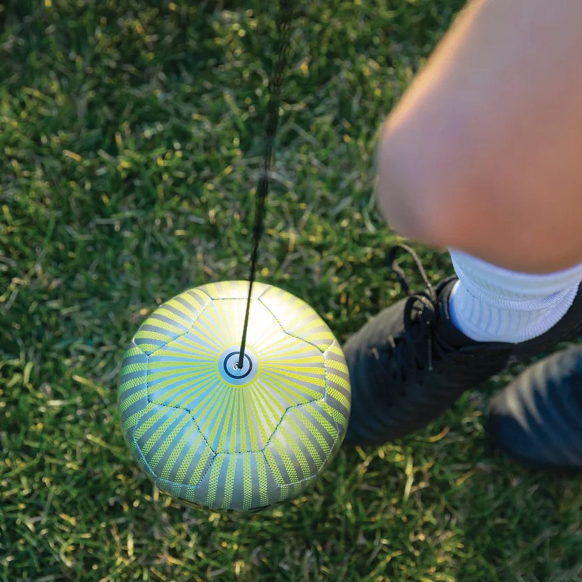 Person stepping on a green and blue striped soccer star kick touch trainer ball with a string attached on grass.