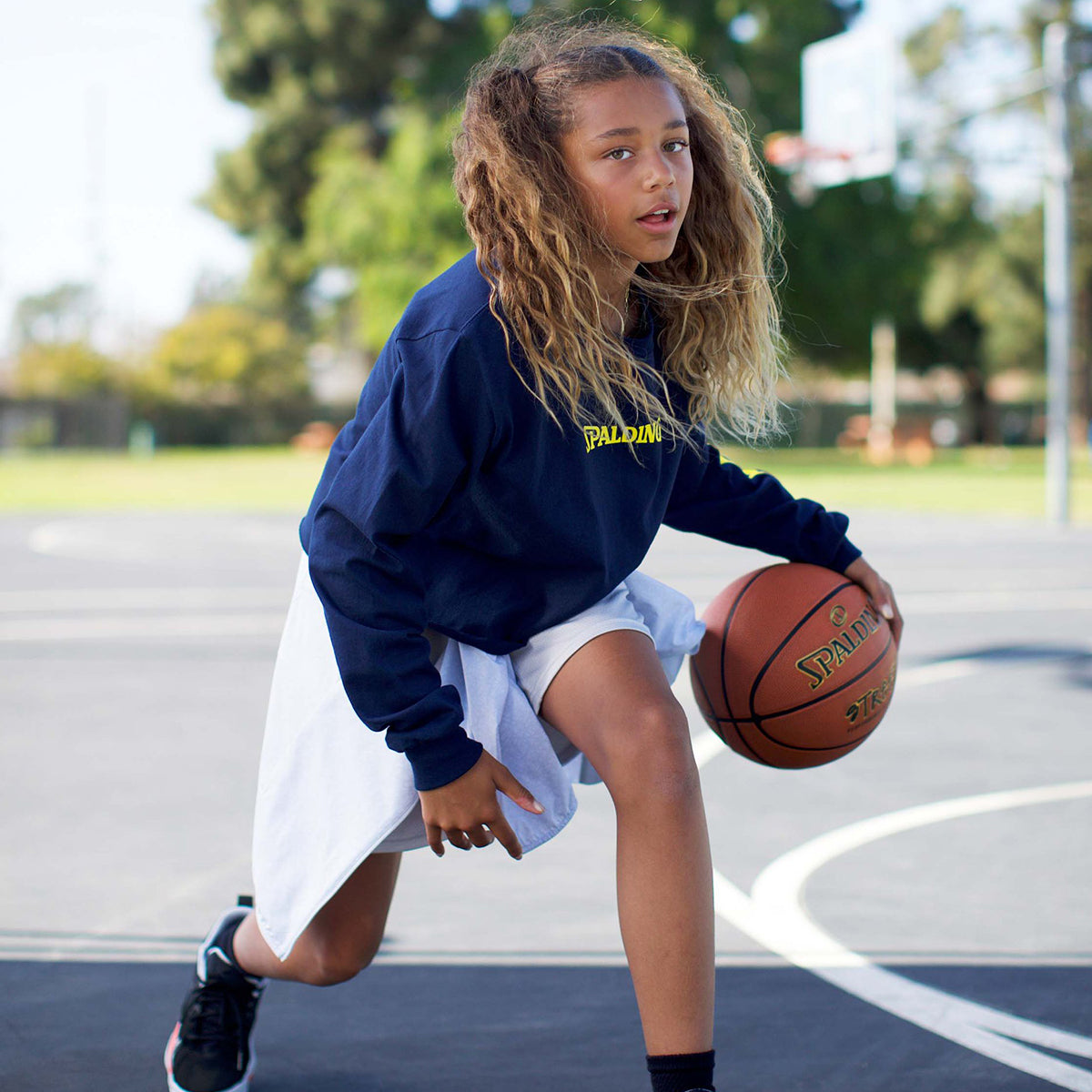 Young girl in a blue sweatshirt and white skirt holding a basketball on a court - Forza Sports