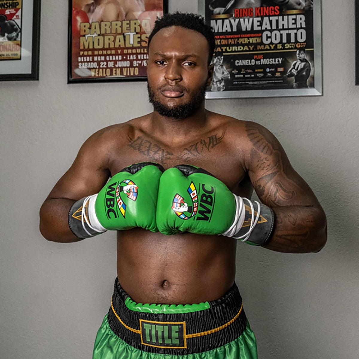 Boxer wearing green boxing gloves and shorts with WBC and Title branding, standing in a room with boxing posters on the wall.