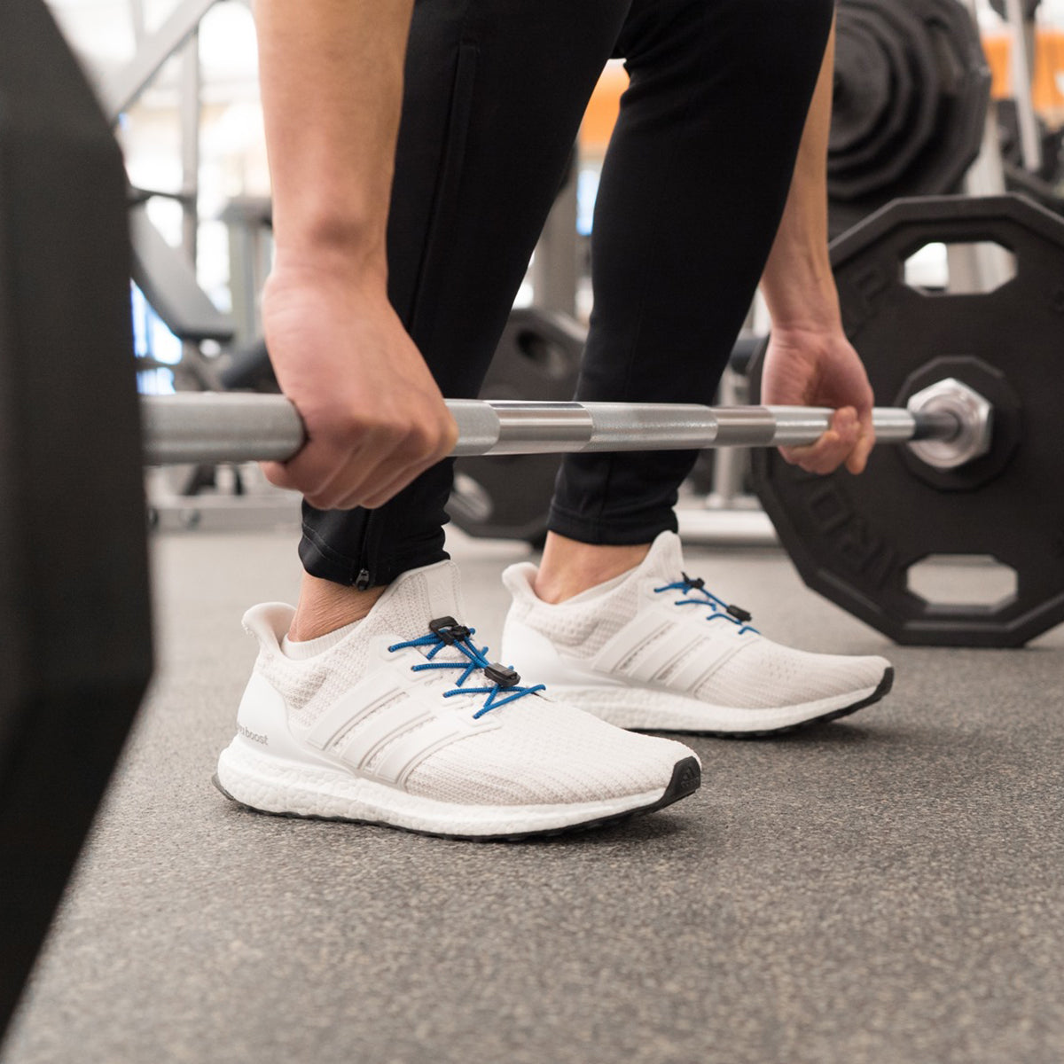 Person lifting a barbell with white sneakers featuring blue laces in a gym setting