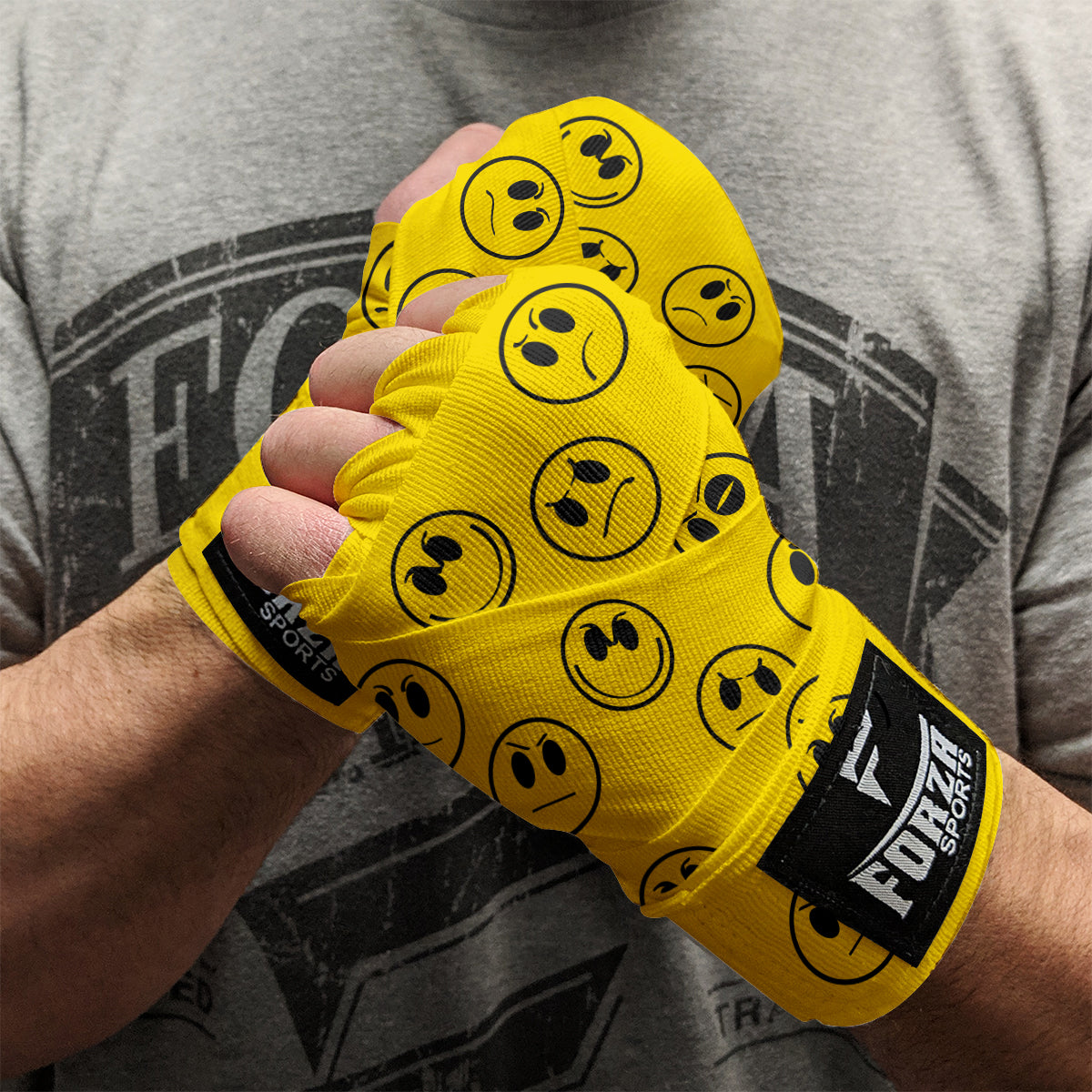 Yellow boxing hand wraps with smiley face patterns worn by a person, against a gray t-shirt background.