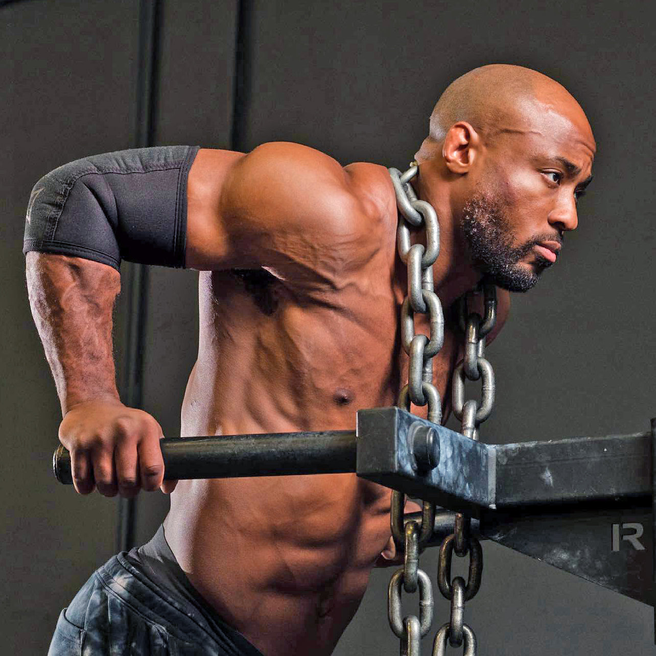 एक गहरे रंग की पृष्ठभूमि पर, जंजीरों से बंधे बारबेल की मदद से एक मांसल व्यक्ति पुश-अप कर रहा है।